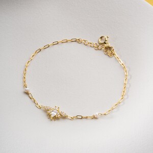 May include: A delicate gold-toned bracelet featuring a bee charm. The bee is adorned with clear crystals and a central pear-shaped stone. The bracelet has a chain link design with small pearl accents.
