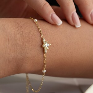 May include: Gold-toned bracelet featuring a bee-shaped pendant adorned with clear crystals and a central pear-shaped stone. The bracelet includes small, round pearls and is displayed on a wrist.