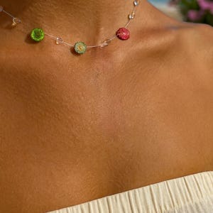 May include: A delicate necklace featuring small, colorful floral accents. The necklace has a transparent string with green, yellow, and red flower-shaped beads. The background shows a blurred view of the ocean and sky.