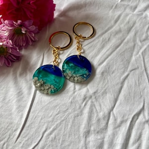 May include: Two circular keyrings with gold-coloured rings and chains. The keyrings feature a blue and teal resin design with small, light-coloured stones. Pink and purple flowers are in the background.