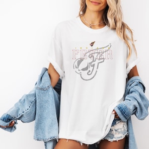 May include: White t-shirt with the text "INDIANA FEVER" in a maroon font and a gray graphic design. The design features a stylized basketball and the letter "F". The shirt has short sleeves.