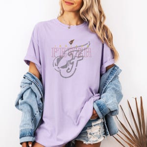 A woman wearing a purple t-shirt with a graphic design and denim jacket stands against a white background.