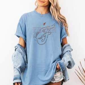 A woman wearing a blue t-shirt with a graphic design and denim jacket stands against a white background.