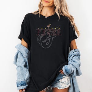 A woman wearing a black t-shirt with the text "INDIE FEAR" printed on it, along with a graphic design. She is also wearing a denim jacket and distressed denim shorts.