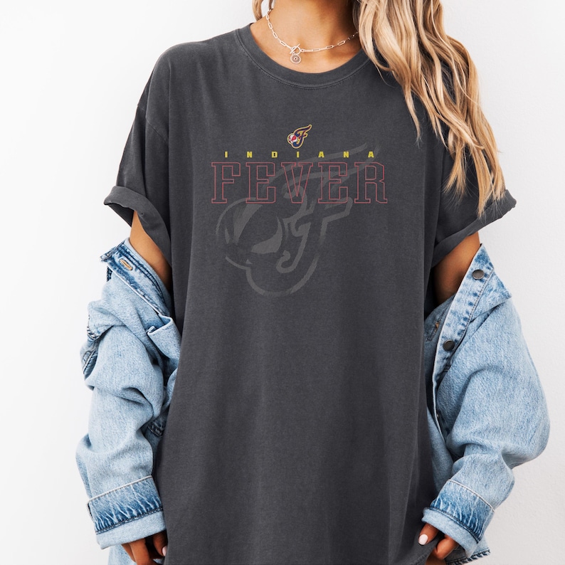 A person wearing a grey t-shirt with the text "INDIANA FEVER" printed on it, along with a graphic design. They are also wearing a denim jacket and have their arms crossed.