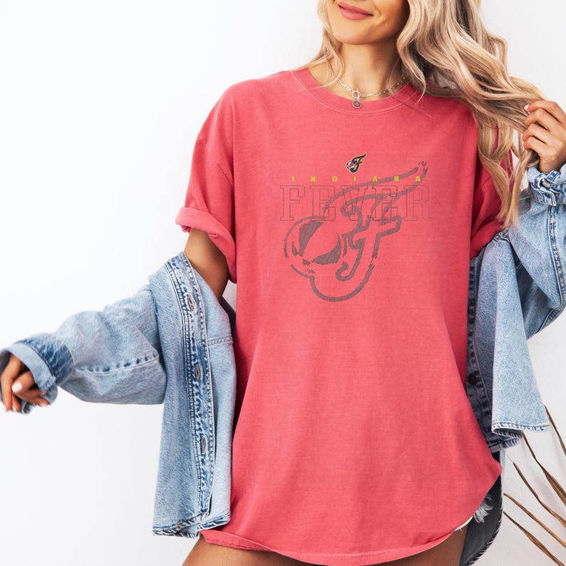 A woman wearing a red t-shirt with a graphic design, a denim jacket, and jeans stands against a white background.