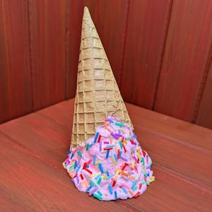 May include: A decorative ice cream cone sculpture. The cone is a light brown color, and the ice cream is pink with colorful sprinkles. The sculpture is set against a reddish-brown wooden background.