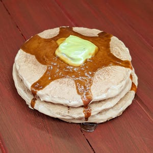 May include: A stack of three pancakes, topped with a pat of green butter and drizzled with syrup. The pancakes are light brown, and the syrup is a darker brown. The image is set against a red wooden background.