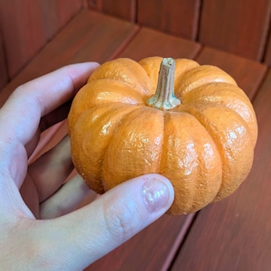 May include: A small, orange pumpkin with a textured surface and a brown stem. The pumpkin is round with visible ridges. It is a decorative item, likely for fall or autumn-themed decor.