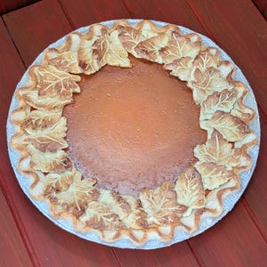 May include: A whole pumpkin pie with a golden-brown crust and a smooth, orange filling. The crust is decorated with leaf-shaped cutouts around the edge. The pie sits on a white plate, ideal for a seasonal celebration.