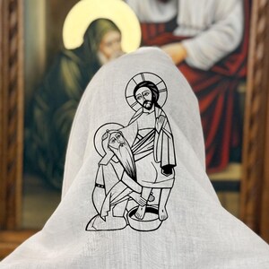 May include: A pale grey hooded garment featuring a black and white line drawing of a religious figure washing another's feet. The background shows a framed painting with religious figures.