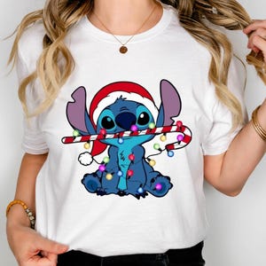 Peut inclure: Un t-shirt blanc à manches courtes avec un motif du personnage bleu Stitch portant un bonnet de Père Noël rouge, tenant une canne en sucre rouge et blanche dans sa bouche et emmêlé dans une guirlande de lumières de Noël colorées.