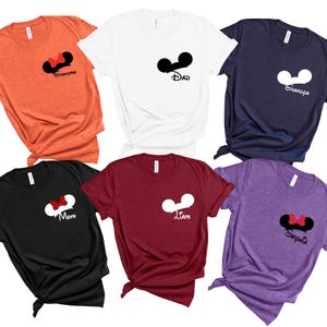 May include: A collection of six t-shirts in various colors, including orange, white, navy, black, burgundy, and purple. Each shirt features a Mickey Mouse ear design with text such as "Grandma," "Dad," "Grandpa," "Mom," "Liam," and "Sophia."