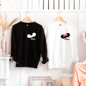May include: Black sweatshirt with white Mickey Mouse ears and the word "Dad" in a script font. A white sweatshirt features Minnie Mouse ears with a red bow and the word "Mom". Both are hanging on wooden hangers.