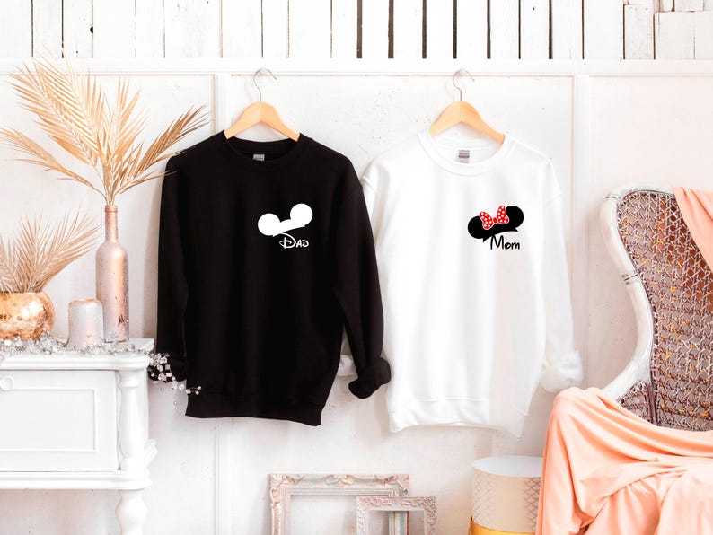 May include: Black sweatshirt with white Mickey Mouse ears and the word "Dad" in a script font. A white sweatshirt features Minnie Mouse ears with a red bow and the word "Mom". Both are hanging on wooden hangers.