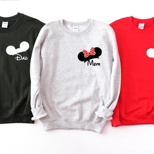 May include: Three sweatshirts in black, gray, and red. Each sweatshirt features a white Mickey Mouse head design. The black sweatshirt says "Dad", the gray sweatshirt has a Minnie Mouse bow and says "Mom", and the red sweatshirt says "Lucas".