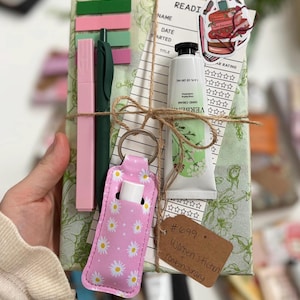May include: A wrapped gift set with green floral paper, twine, and a pink daisy-patterned lip balm holder. It includes a pink highlighter, green pen, hand cream, a bookmark, and a tag that reads "Women's Fiction Contemporary".