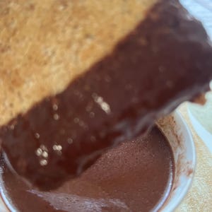 May include: A close-up shot of a piece of toasted bread dipped into a cup of rich, dark chocolate sauce. The bread is golden brown, and the chocolate is a deep, glossy brown. The cup is white.