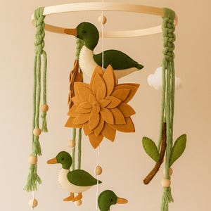 May include: A handcrafted baby mobile with green and white felt ducks, a large mustard-coloured flower, and a wooden ring. The mobile includes green macrame accents, wooden beads, and a small white cloud. The ducks have orange beaks and feet.
