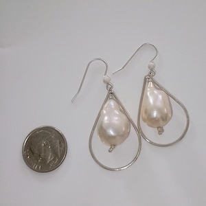 Puede incluir: Un par de pendientes plateados con forma de lágrima y grandes perlas barrocas blanquecinas. Los pendientes tienen un diseño sencillo y elegante, con las perlas suspendidas dentro de los marcos plateados. Una moneda está al lado de los pendientes para comparar el tamaño.