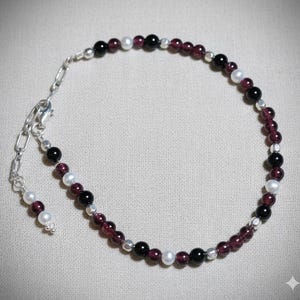 Op de afbeelding: Een delicate armband met een mix van kleine, ronde kralen. De armband combineert dieprode granaat kralen, zwarte kralen en witte parel kralen, afgewisseld met kleine zilverkleurige kralen. De sluiting en ketting zijn ook zilverkleurig.