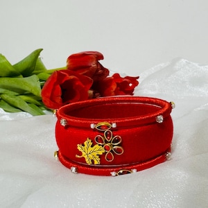 Può includere: Due braccialetti rossi con dettagli dorati e strass. Un braccialetto presenta un design a foglia d'acero e fiore dorato con gemme rosse. I braccialetti sono su uno sfondo bianco con fiori rossi.