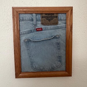 May include: A light blue denim fabric section from Wrangler jeans, featuring a brown leather patch with the text Wrangler Premium Quality and a small red Wrangler tag, displayed inside a rectangular wooden frame with a natural grain finish.