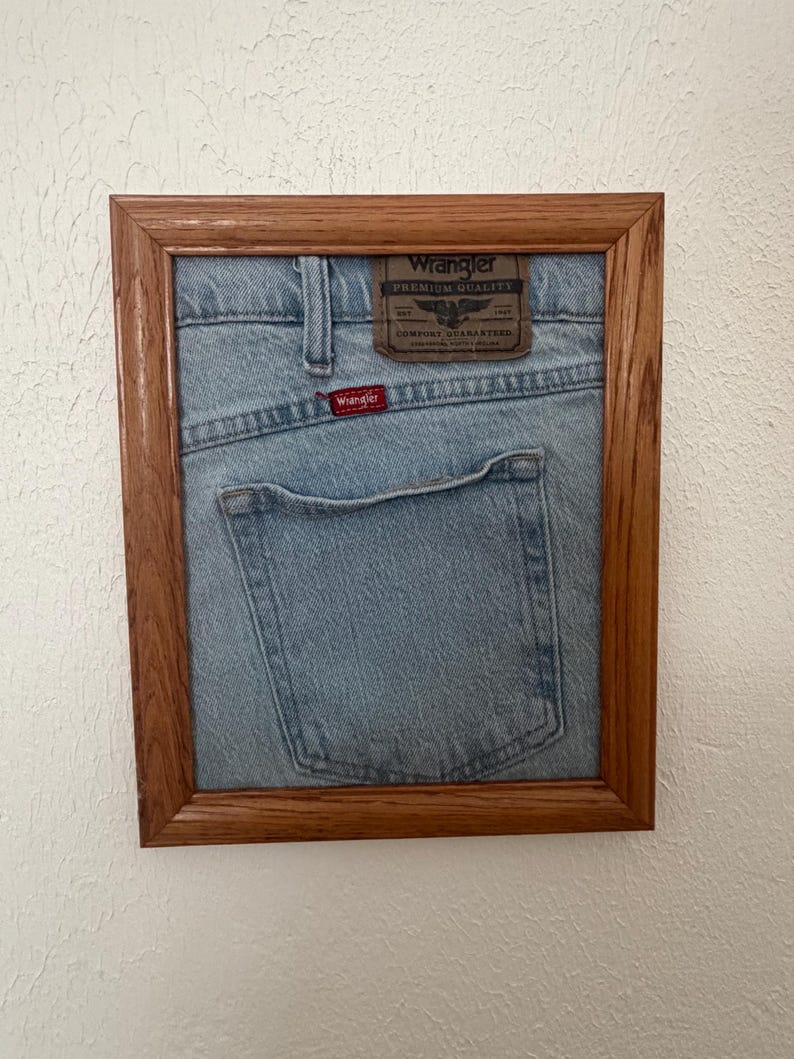 May include: A light blue denim fabric section from Wrangler jeans, featuring a brown leather patch with the text Wrangler Premium Quality and a small red Wrangler tag, displayed inside a rectangular wooden frame with a natural grain finish.