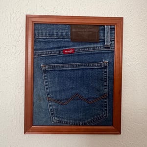 May include: A framed piece of blue denim fabric featuring the back pocket of a pair of Wrangler jeans. The denim includes a brown leather patch at the top, a red Wrangler brand tag, and the signature W stitching on the back pocket, all encased in a wooden frame.
