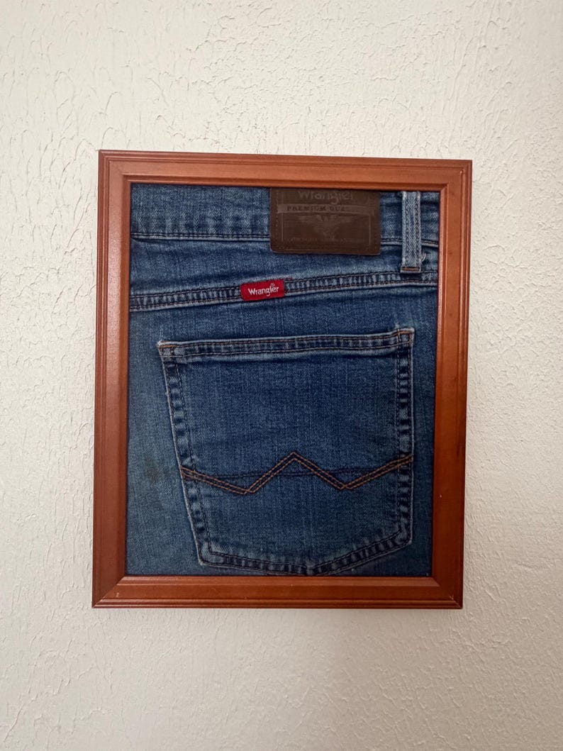 May include: A framed piece of blue denim fabric featuring the back pocket of a pair of Wrangler jeans. The denim includes a brown leather patch at the top, a red Wrangler brand tag, and the signature W stitching on the back pocket, all encased in a wooden frame.