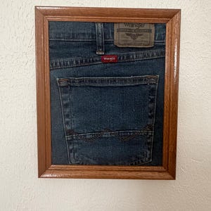 May include: A framed piece of dark blue denim fabric featuring a Wrangler back pocket, a brown leather-look patch with the text Wrangler Premium Quality, and a small red Wrangler brand tag, all encased in a simple rectangular wooden frame.