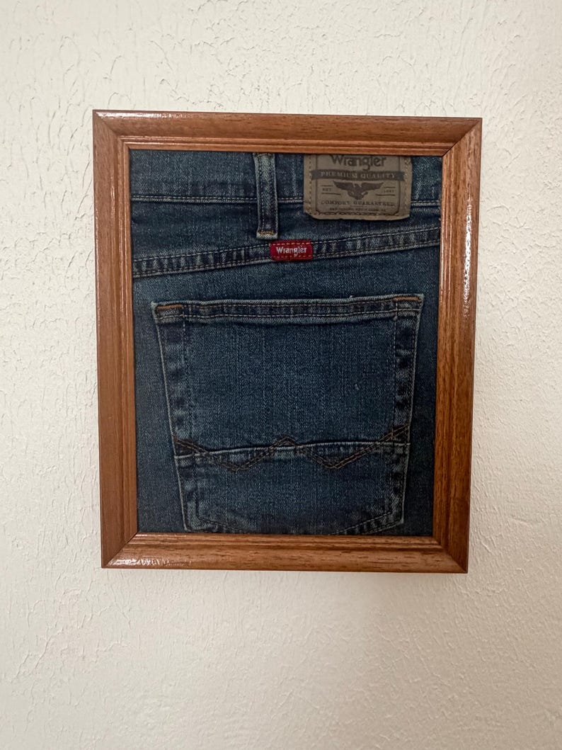 May include: A framed piece of dark blue denim fabric featuring a Wrangler back pocket, a brown leather-look patch with the text Wrangler Premium Quality, and a small red Wrangler brand tag, all encased in a simple rectangular wooden frame.