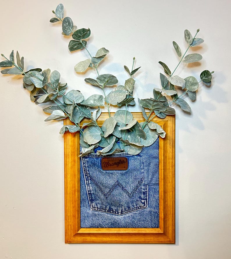 May include: A rectangular wooden picture frame containing a blue denim fabric panel with a Wrangler pocket featuring the brand logo. Sprigs of green eucalyptus leaves extend upward from the top of the denim pocket and out of the frame.