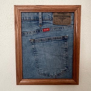 May include: A framed piece of blue denim fabric featuring the back pocket of a pair of Wrangler jeans. The fabric includes a brown leather-like patch with the text Wrangler Premium Quality and a small red tag with the Wrangler logo.