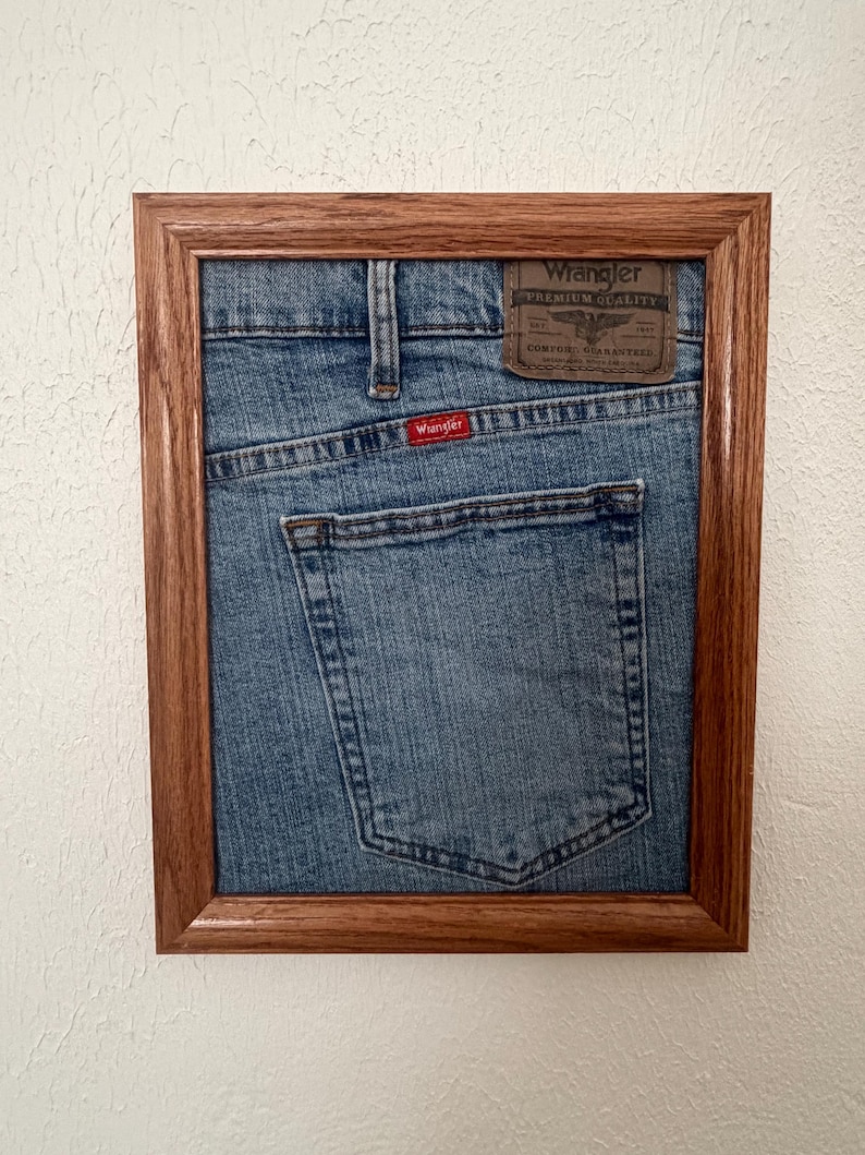 May include: A framed piece of blue denim fabric featuring the back pocket of a pair of Wrangler jeans. The fabric includes a brown leather-like patch with the text Wrangler Premium Quality and a small red tag with the Wrangler logo.