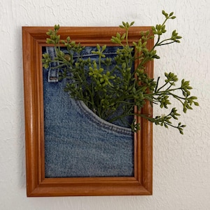 May include: A framed artwork with a denim pocket and green sprigs. The wooden frame is brown, and the denim pocket is light blue. The green sprigs provide a natural contrast to the denim and wooden frame.