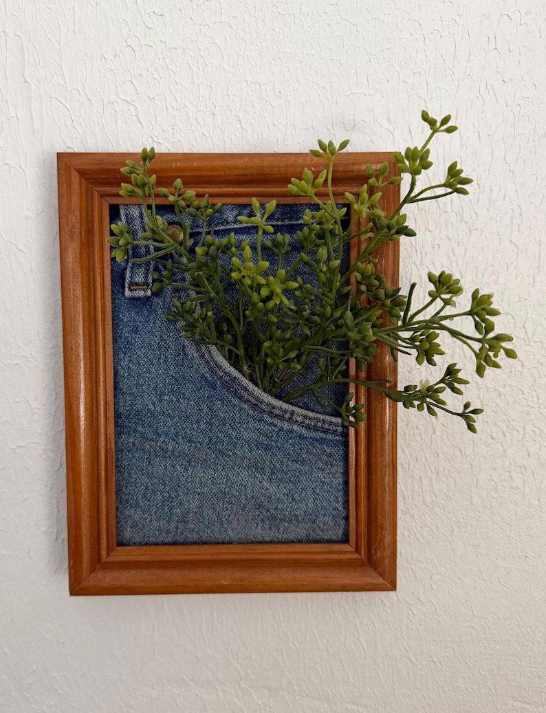 May include: A framed artwork with a denim pocket and green sprigs. The wooden frame is brown, and the denim pocket is light blue. The green sprigs provide a natural contrast to the denim and wooden frame.