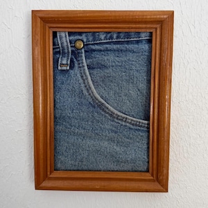 May include: A framed artwork showing a close-up of a blue denim jeans pocket. The pocket has detailed stitching and a brass button. The wooden frame has a warm, brown finish, giving a rustic appearance.