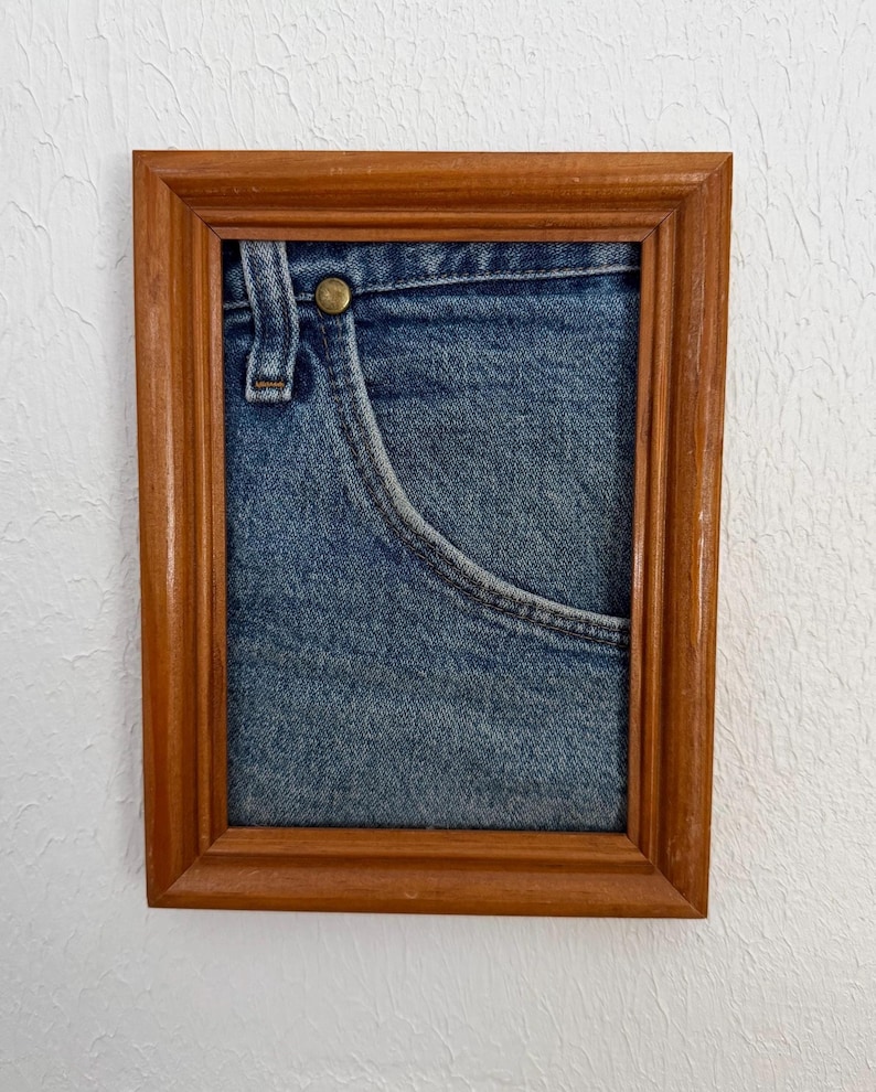 May include: A framed artwork showing a close-up of a blue denim jeans pocket. The pocket has detailed stitching and a brass button. The wooden frame has a warm, brown finish, giving a rustic appearance.