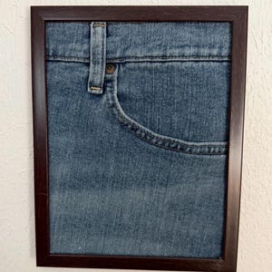 May include: A framed artwork showing a close-up of a blue denim jeans pocket and waistband. The frame is dark brown. The denim has visible stitching and a button detail. The artwork is mounted on a white wall.