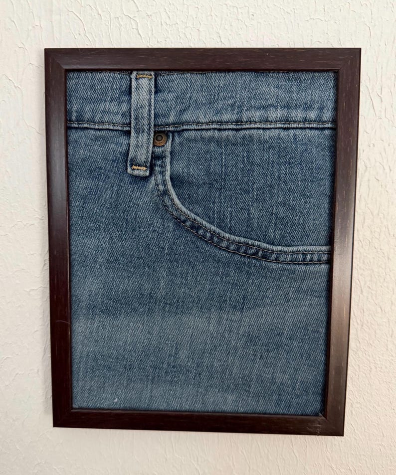May include: A framed artwork showing a close-up of a blue denim jeans pocket and waistband. The frame is dark brown. The denim has visible stitching and a button detail. The artwork is mounted on a white wall.