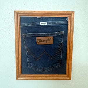 May include: A framed piece of dark blue denim fabric featuring a Wrangler back pocket with a brown leather patch and a white Wrangler brand tag. The denim is mounted inside a rectangular wooden frame with a natural light brown finish.