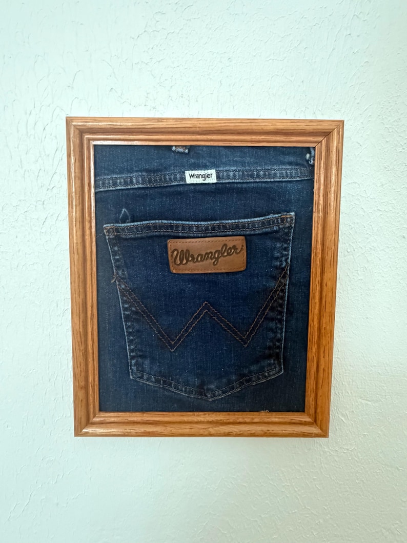 May include: A framed piece of dark blue denim fabric featuring a Wrangler back pocket with a brown leather patch and a white Wrangler brand tag. The denim is mounted inside a rectangular wooden frame with a natural light brown finish.