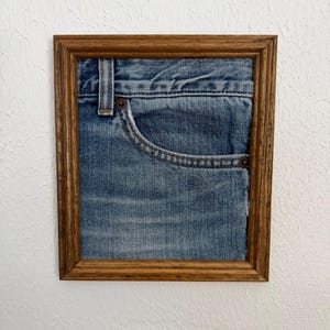 May include: A framed artwork featuring a section of blue denim jeans. The jeans are displayed within a rectangular wooden frame, showcasing the pocket and stitching details. The artwork is mounted on a white textured wall.