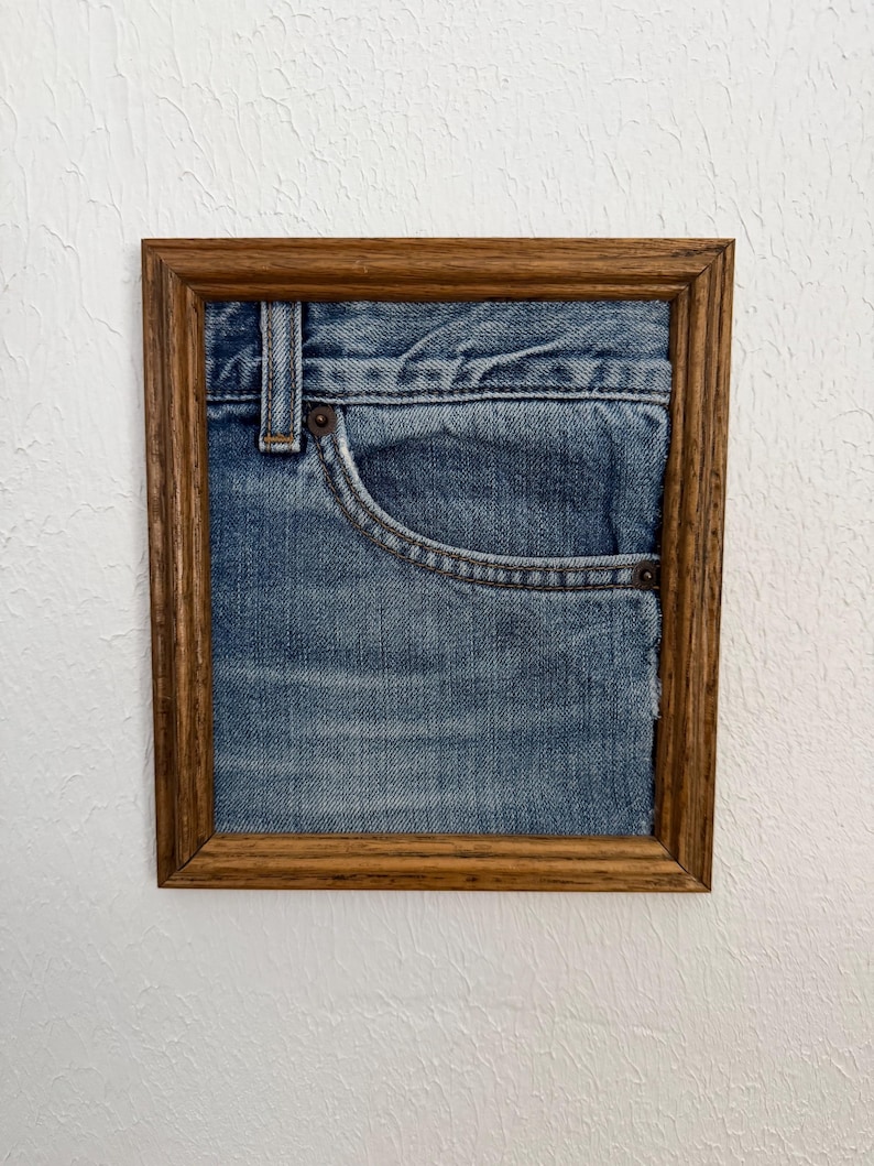 May include: A framed artwork featuring a section of blue denim jeans. The jeans are displayed within a rectangular wooden frame, showcasing the pocket and stitching details. The artwork is mounted on a white textured wall.