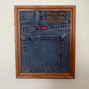 May include: A framed piece of blue denim fabric featuring the back pocket of a pair of Wrangler jeans. The denim includes a brown leather-like patch with the text Wrangler Premium Quality Comfort Guaranteed and a small red tag with the word Wrangler.