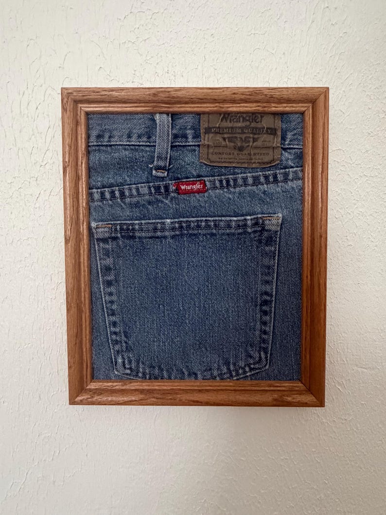 May include: A framed piece of blue denim fabric featuring the back pocket of a pair of Wrangler jeans. The denim includes a brown leather-like patch with the text Wrangler Premium Quality Comfort Guaranteed and a small red tag with the word Wrangler.