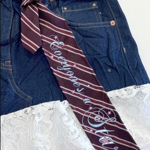 Puede incluir: Falda vaquera azul oscuro con cinturón de corbata a rayas burdeos y blancas. La corbata presenta el texto "Everyone's a Star" en cursiva con detalles de estrellas. La falda tiene un ribete de encaje blanco.