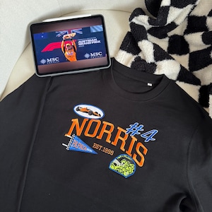 May include: Black sweatshirt featuring "NORRIS" in orange and blue embroidery, with the number "4" and racing patches. A tablet displays a race event. This is a casual item of clothing.