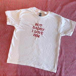 Puede incluir: Camiseta blanca con el texto rojo bordado "BUT DADDY I LOVE HIM". La camiseta es de manga corta y está hecha de un material suave. Se muestra sobre una superficie a cuadros rojos y blancos.